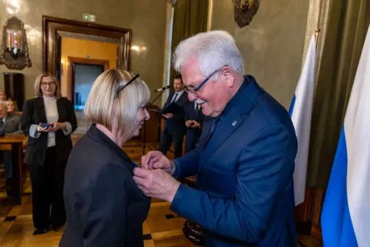 Wręczenie odznaczenia przez mężczyznę kobiecie w eleganckich strojach. W tle flagi i osoby obserwujące ceremonię.
