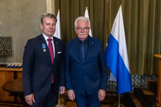 Dwaj mężczyźni w garniturach stoją obok siebie, w tle flagi.