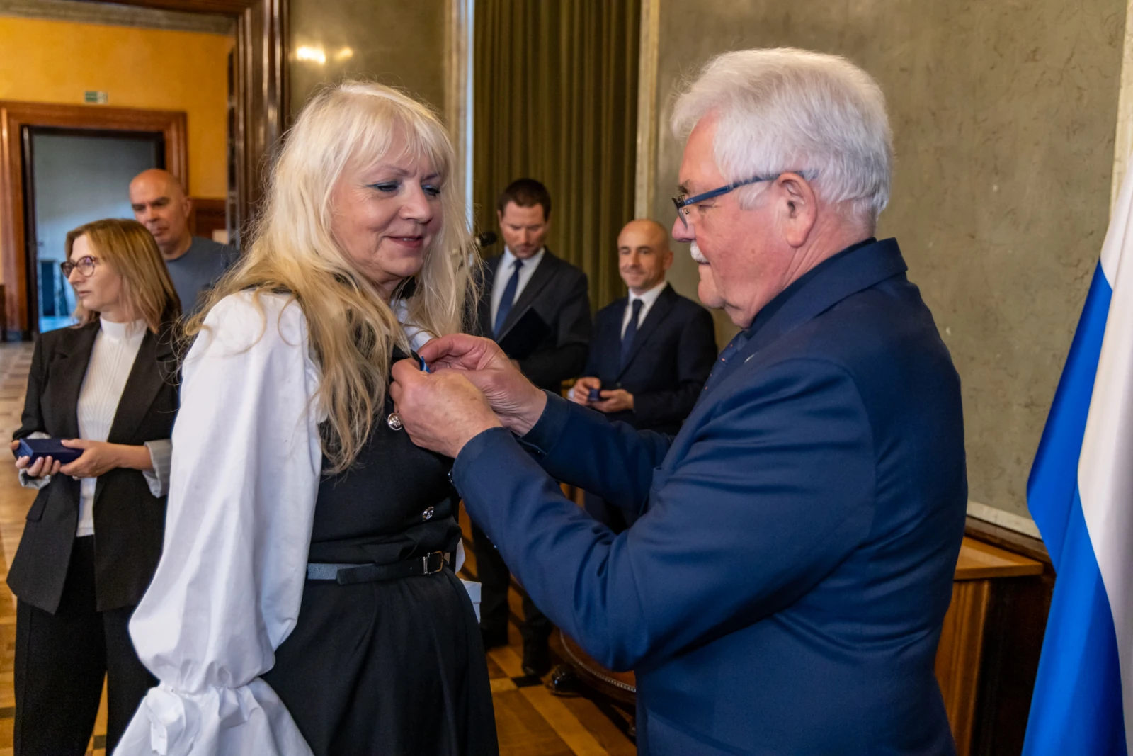 Wręczenie odznaczenia kobiecie przez mężczyznę w formalnym otoczeniu.  Autor: Jan Graczyński