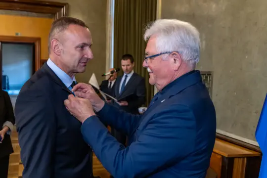 Mężczyzna odbierający odznaczenie od innego mężczyzny w formalnym otoczeniu.