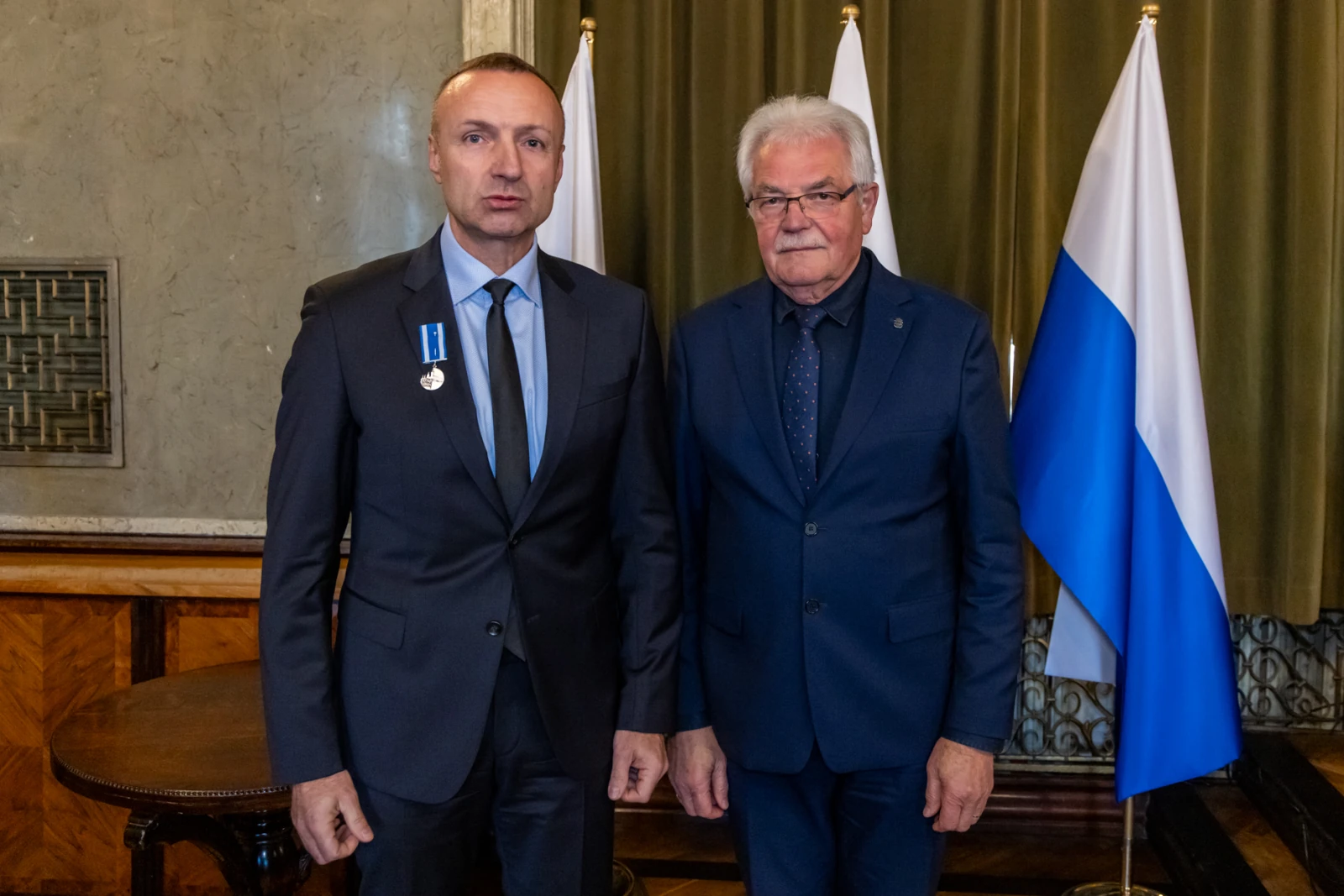 Dwaj mężczyźni w garniturach, jeden z medalem, stoją obok flag.  Autor: Jan Graczyński