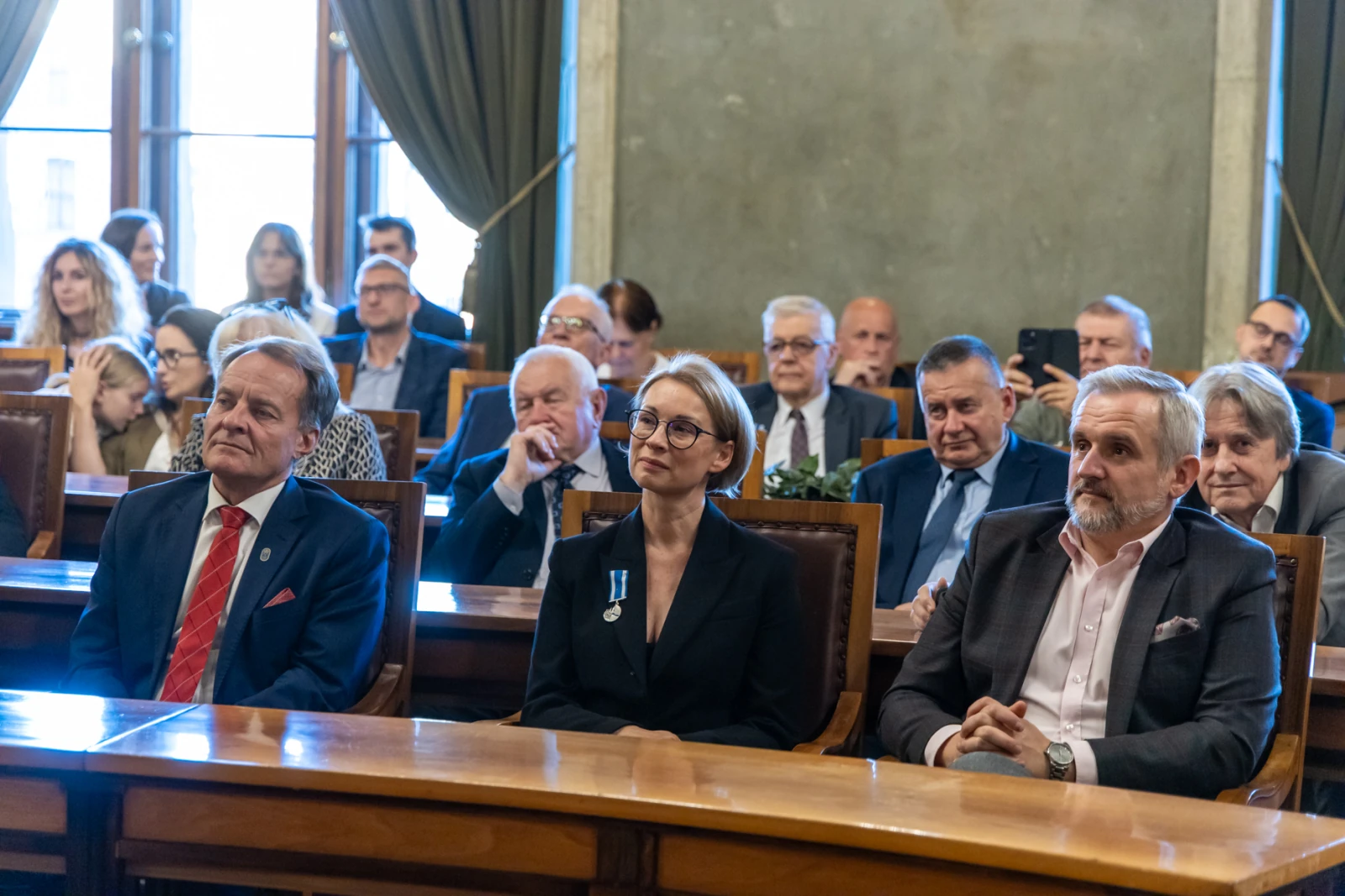 Uczestnicy uroczystości w sali, niektórzy z odznaczeniami, wśród nich kobieta w okularach.  Autor: Jan Graczyński