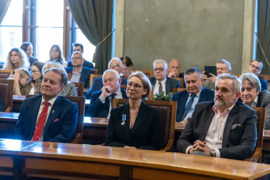 Uczestnicy uroczystości w sali, niektórzy z odznaczeniami, wśród nich kobieta w okularach.