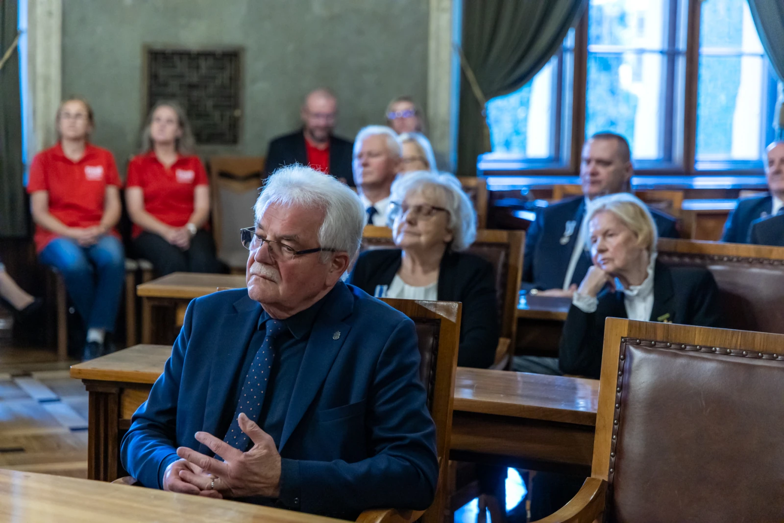 Mężczyzna w garniturze siedzi w sali obrad, w tle grupa osób w czerwonych koszulkach.  Autor: Jan Graczyński