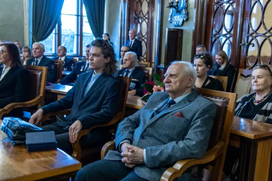 Grupa osób w formalnych strojach siedzi w sali, w tle okna i dekoracyjne elementy architektoniczne.