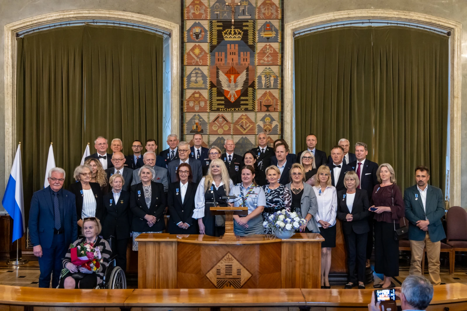 Grupa ludzi w formalnych strojach, stojących w sali z herbem i flagami.  Autor: Jan Graczyński