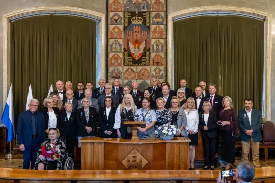 Grupa ludzi w formalnych strojach, stojących w sali z herbem i flagami.