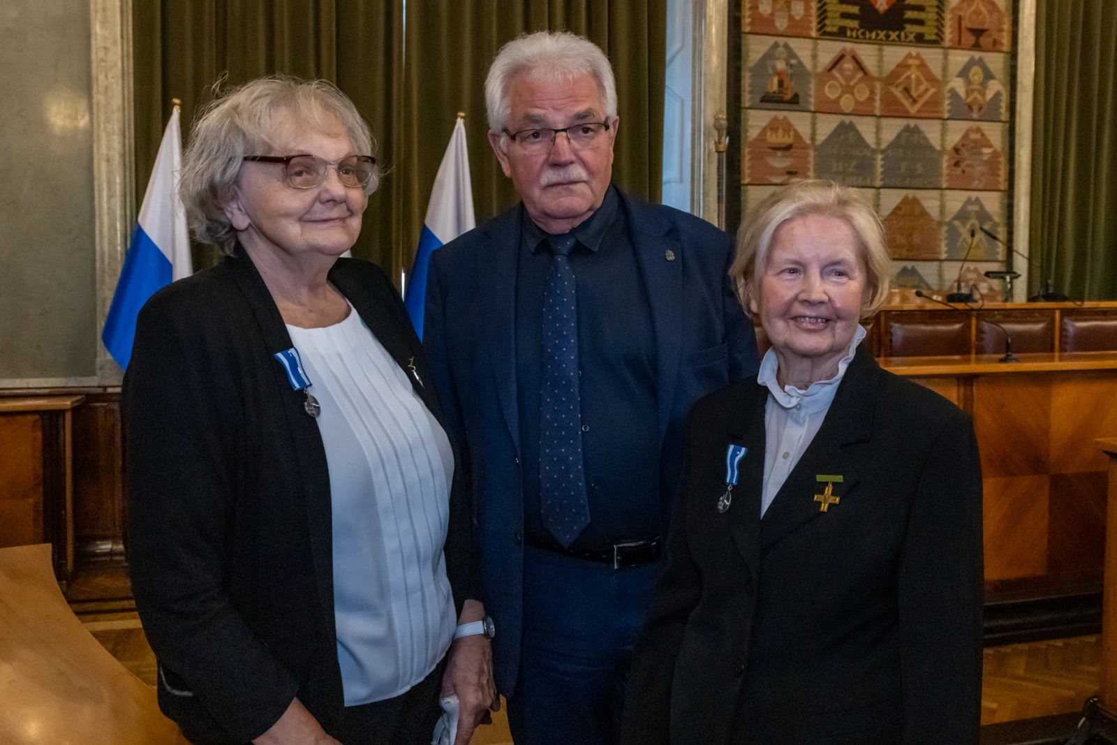 Trzy osoby w formalnych strojach, z odznaczeniami, w sali z flagami.  Autor: Jan Graczyński