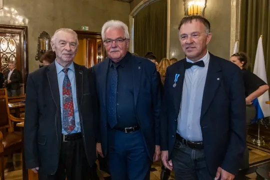 Trzech mężczyzn w eleganckich strojach, stojących obok siebie w stylowym wnętrzu.