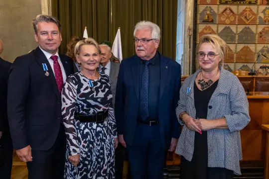 Cztery osoby w formalnych strojach, uśmiechające się, w tle sala z flagami.