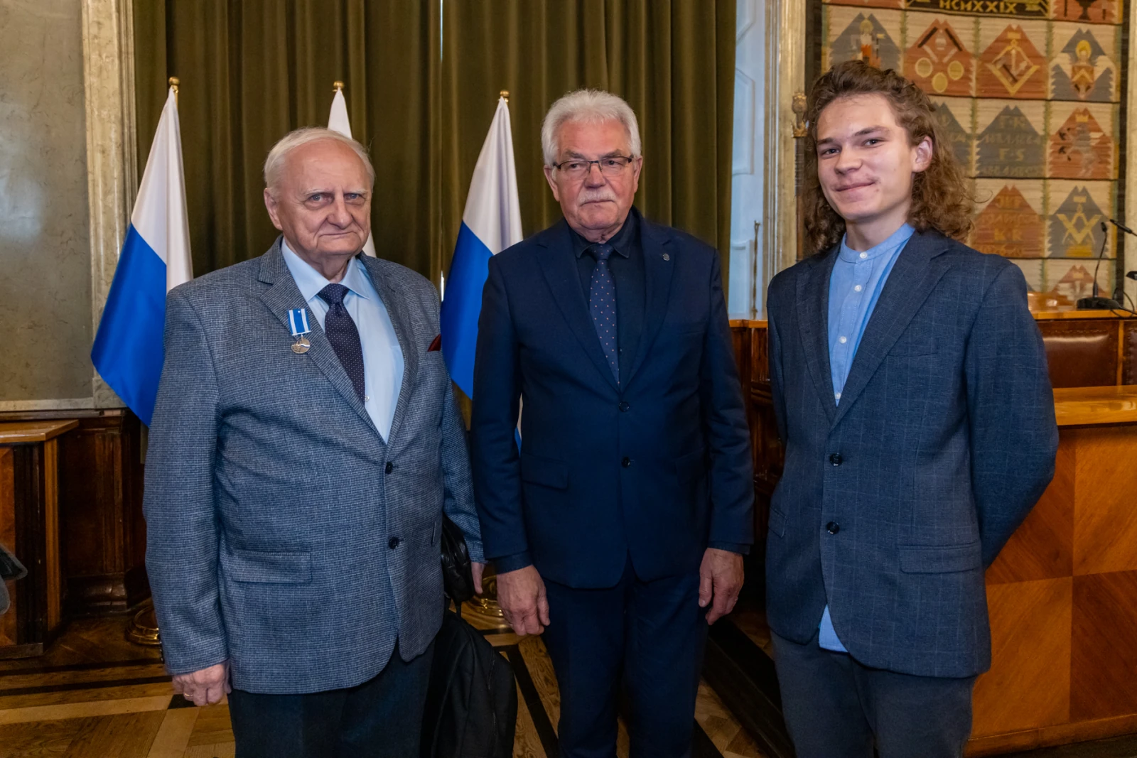 Trzech mężczyzn w formalnych strojach, stojących obok siebie w sali z flagami.  Autor: Jan Graczyński