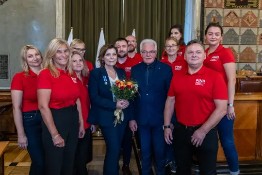 Grupa osób w czerwonych koszulkach, wręczająca bukiet kwiatów w uroczystym otoczeniu.