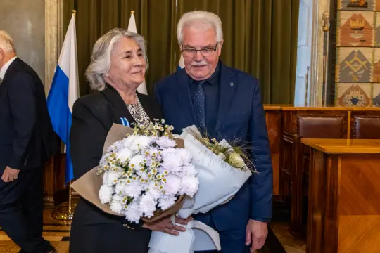 Para osób trzymająca bukiety kwiatów, w tle flagi i eleganckie wnętrze.
