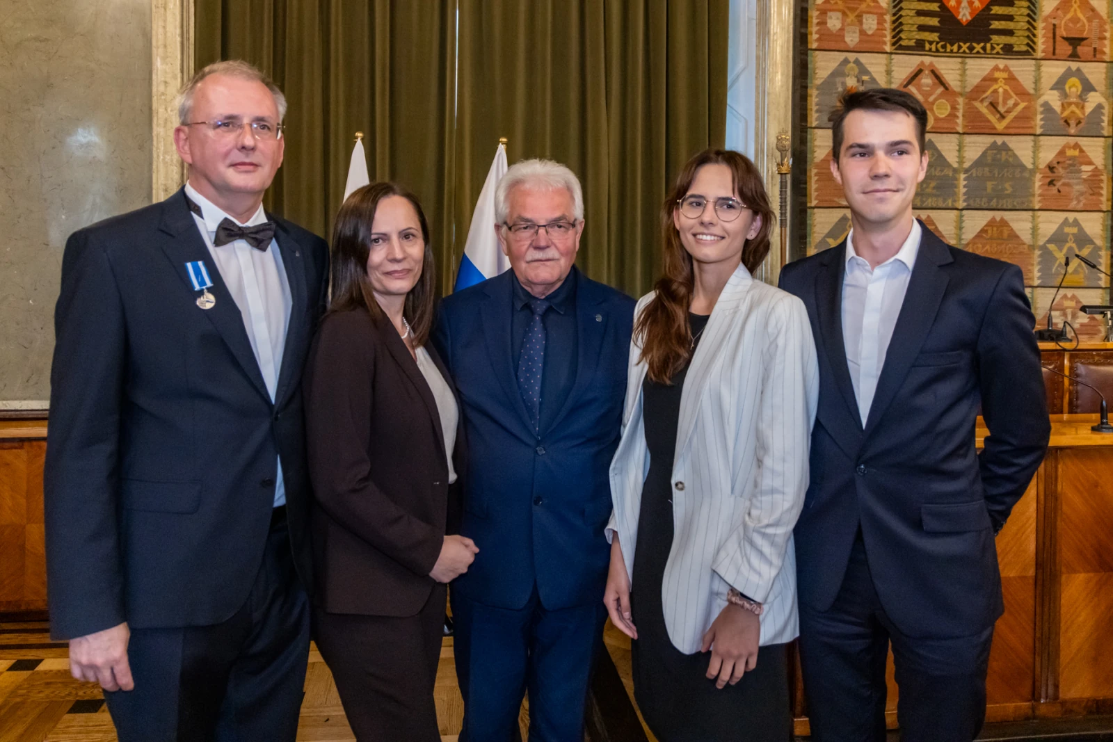 Grupa pięciu osób w formalnych strojach, stojących w sali z flagami w tle.  Autor: Jan Graczyński