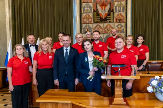 Grupa ludzi w czerwonych koszulkach, z kwiatami i medalami, w sali z dekoracjami.