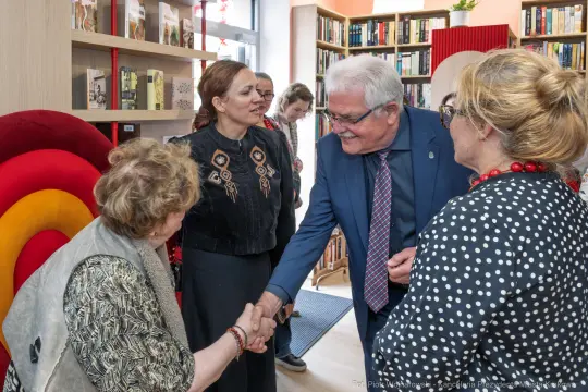 Spotkanie osób w bibliotece, uściski dłoni i rozmowy.
