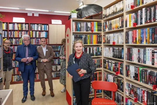Kobieta z teczką w bibliotece, w tle mężczyźni i regały z książkami.