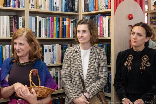 Trzy kobiety w bibliotece, jedna z koszykiem, pozostałe w eleganckich strojach.