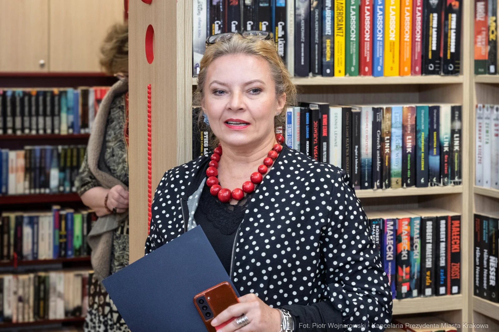 Kobieta w kropkowanej bluzce z czerwonymi koralikami, trzymająca dokumenty w bibliotece.  Autor: Piotr Wojnarowski