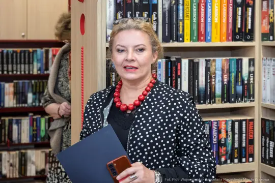 Kobieta w kropkowanej bluzce z czerwonymi koralikami, trzymająca dokumenty w bibliotece.