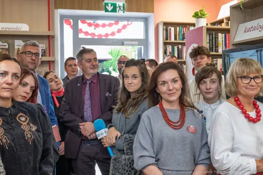 Grupa ludzi w bibliotece, niektórzy z mikrofonem, inni w rozmowie.