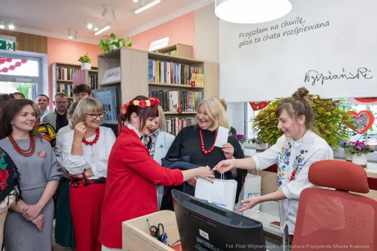 Kobieta w czerwonym stroju wręcza torbę z prezentem, wokół uśmiechnięte osoby w bibliotece.