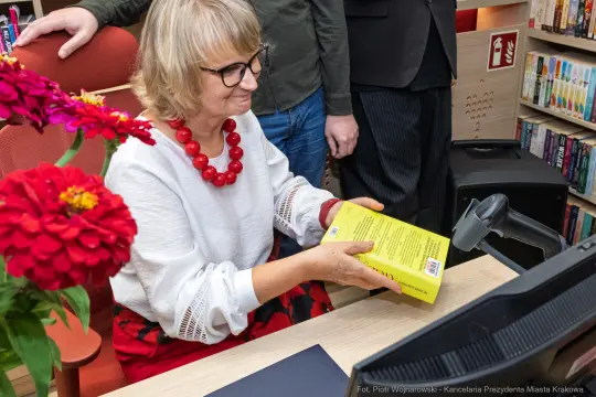 Kobieta z okularami trzyma książkę, siedząc przy biurku w bibliotece. W tle kwiaty i regał z książkami.