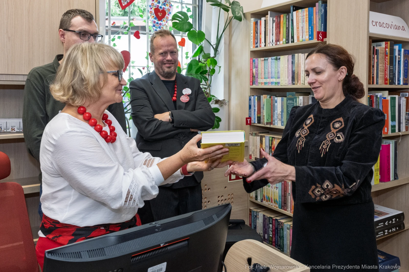Wręczenie książki w bibliotece, uczestnicy w strojach formalnych, dekoracje sercowe w tle.  Autor: Piotr Wojnarowski