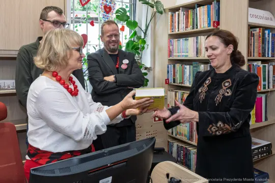 Wręczenie książki w bibliotece, uczestnicy w strojach formalnych, dekoracje sercowe w tle.