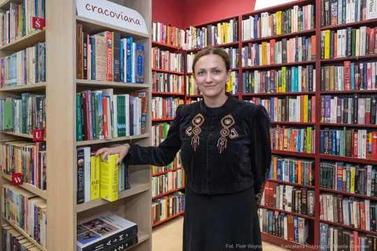 Kobieta w czarnym stroju stoi obok regału z książkami w bibliotece.