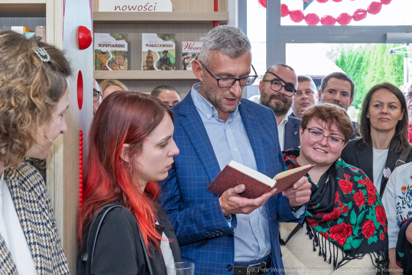 Mężczyzna czyta książkę, wokół niego zebrani ludzie, w tle półki z książkami.  Autor: Piotr Wojnarowski