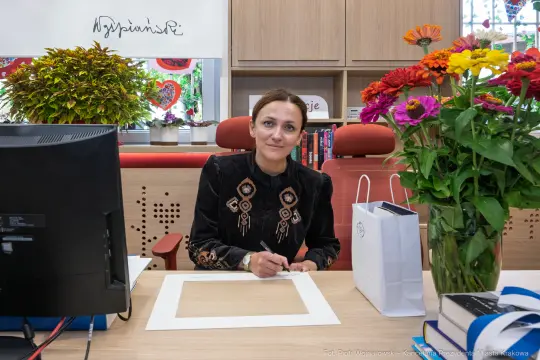 Kobieta w czarnej bluzce podpisuje dokumenty przy biurku z kwiatami i komputerem.