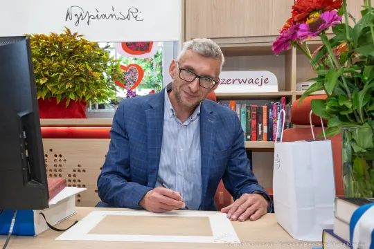 Mężczyzna piszący w biurze, otoczony książkami i kwiatami.