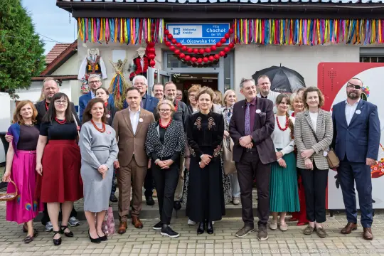 Grupa ludzi w strojach formalnych przed budynkiem, udekorowanym kolorowymi ozdobami.