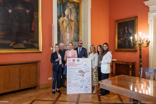 Grupa osób stojąca obok plakatu promującego inicjatywę społeczną w eleganckim wnętrzu.
