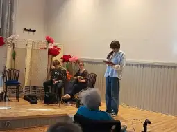 Młoda osoba recytuje tekst, obok niej grają na instrumentach dwie inne osoby.
