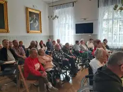 Grupa osób starszych siedzących w sali, część na wózkach inwalidzkich.