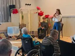 Młoda kobieta śpiewa na scenie, a publiczność w wózkach inwalidzkich słucha jej występu.