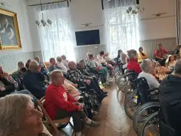 Grupa seniorów siedzących w sali, część z nich na wózkach inwalidzkich.