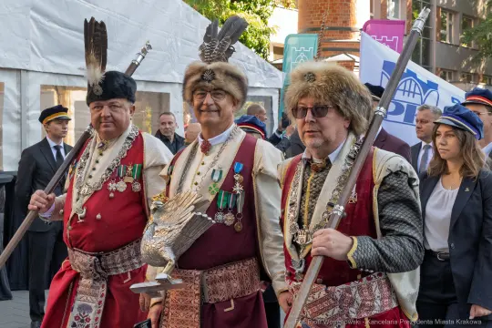 Trzej mężczyźni w tradycyjnych strojach z ozdobami i medalami, z orłem w dłoni.
