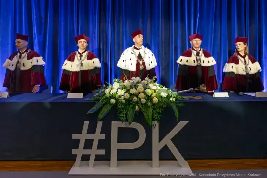 Ceremonia z udziałem osób w togach akademickich przy stole z kwiatami i napisem #PK.