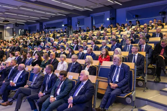 Zgromadzenie ludzi w sali konferencyjnej, większość w garniturach, niektórzy w mundurach.