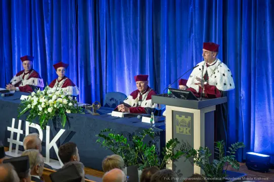 Ceremonia akademicka z mówcą i uczestnikami w togach, dekoracje kwiatowe na stole.