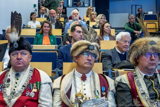 Trzej mężczyźni w tradycyjnych strojach z piórami, siedzący wśród publiczności na wydarzeniu.
