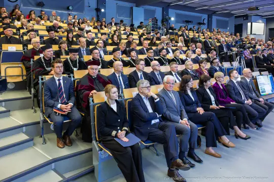 Uczestnicy ceremonii akademickiej siedzą w auli, wśród nich wykładowcy i studenci.