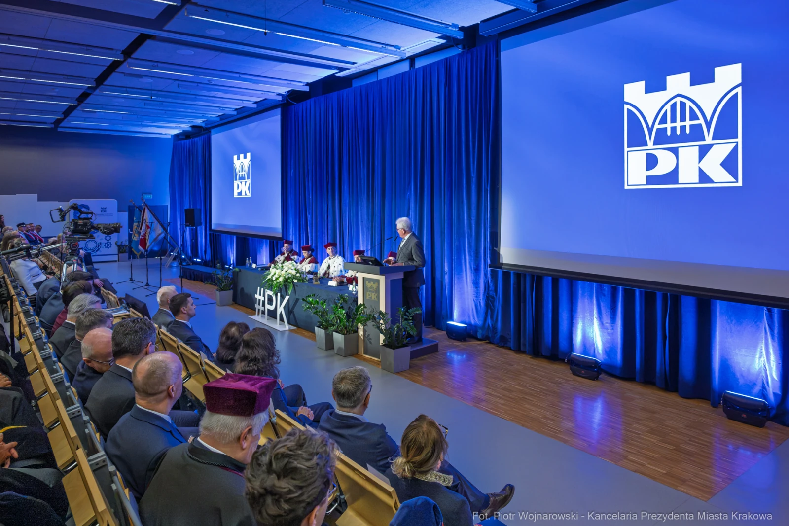 Konferencja z logo PK, prelegent przy mównicy, widownia i flagi.  Autor: Piotr Wojnarowski