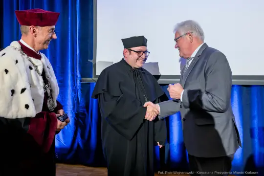 Uroczystość wręczenia nagrody, mężczyźni w togach akademickich i garniturach, uściski dłoni.