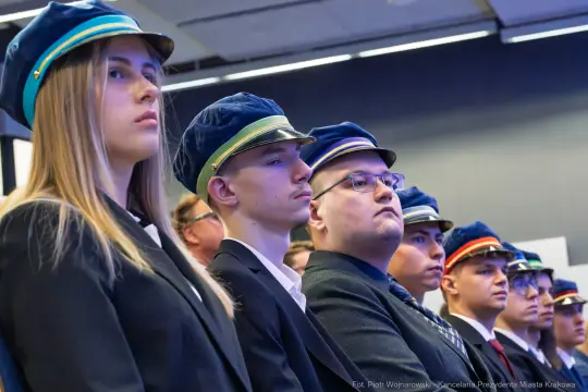 Grupa młodych ludzi w garniturach i czapkach, skupionych na wydarzeniu.