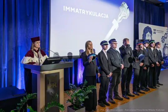 Ceremonia immatrykulacji z przemówieniem i grupą studentów w togach akademickich.