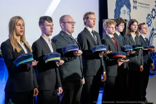 Uczniowie w garniturach trzymają czapki, stojąc w rzędzie na ceremonii.
